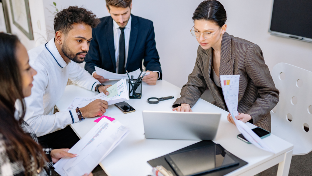 A close-up photograph of a finance leader on a boardroom table reviewing documents, surrounded by the attentive faces of a team seeking direction.